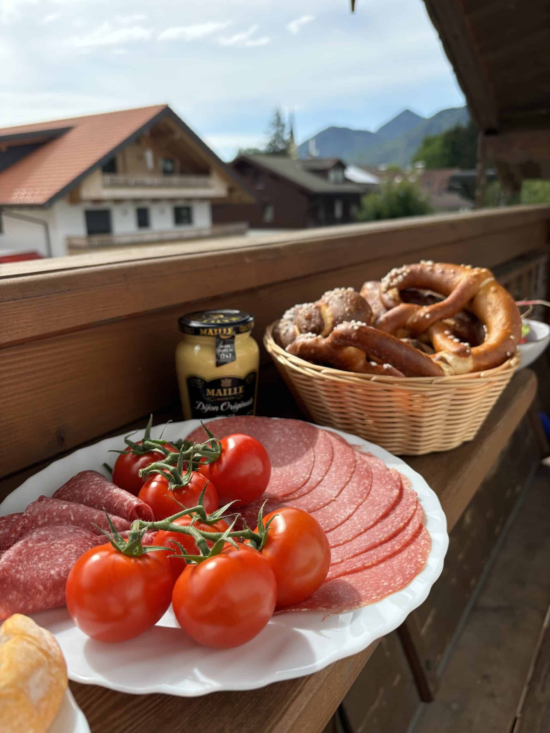 Tomatoes, salami, pretzels on balcony with mountain view.