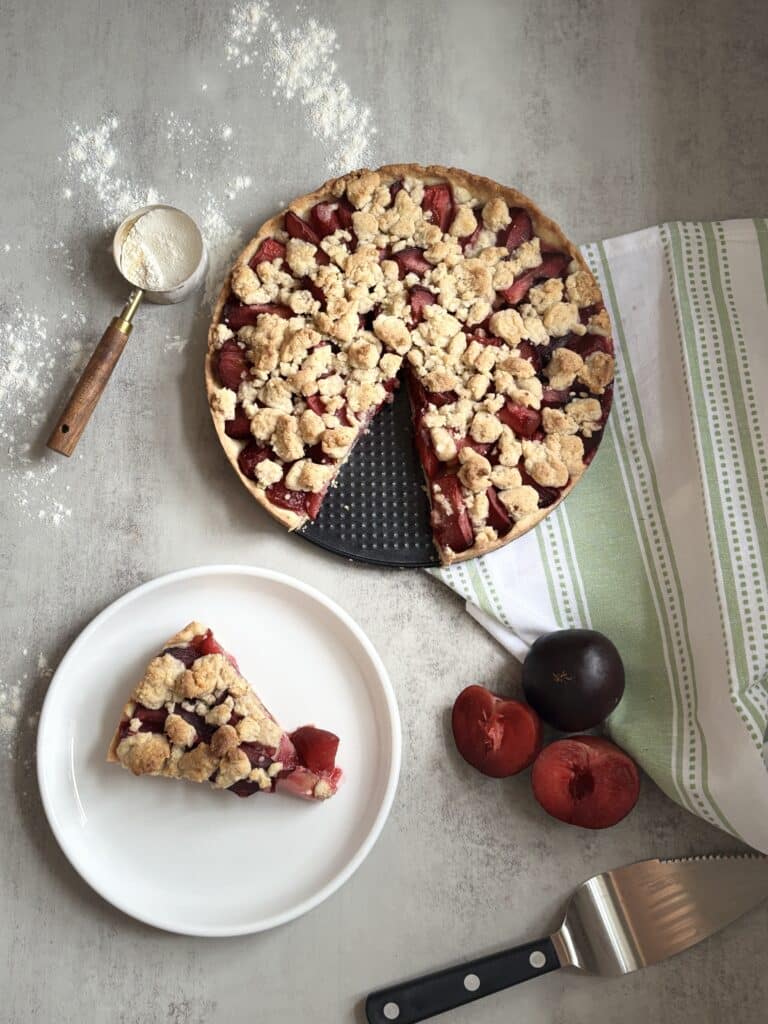 Slice of German plum cake with streusel topping on a plate beside the whole Zwetschgenkuchen