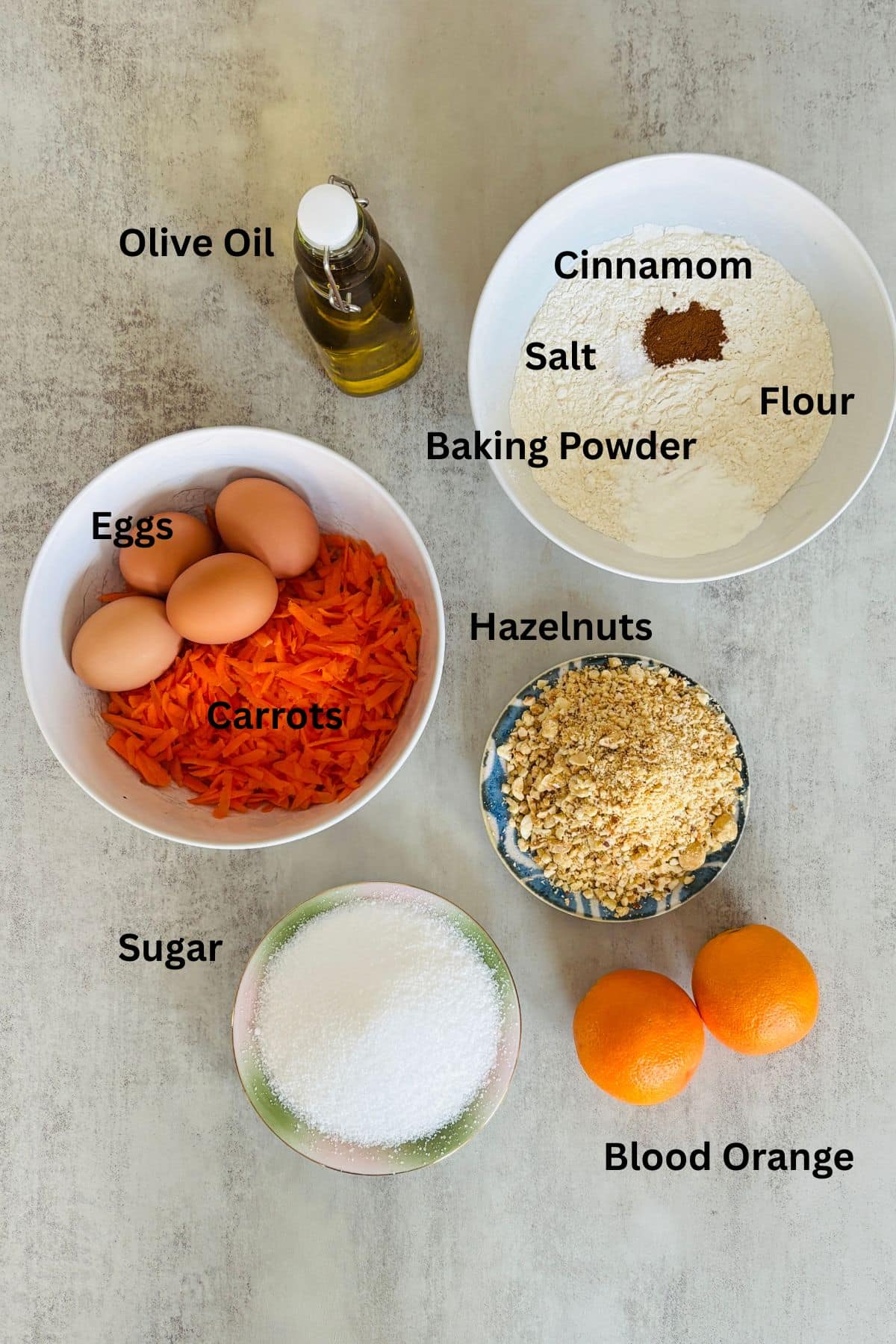 Ingredients for carrot orange cake including shredded carrots, blood oranges, olive oil, hazelnuts, eggs, flour, and sugar arranged on a table
